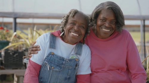 Two women sitting close together outdoors, smiling warmly at the camera. One woman wears a white t-shirt with overalls, and the other wears a pink long-sleeve shirt. They appear to be in a greenhouse or garden setting, with plants and sunlight visible in the background.